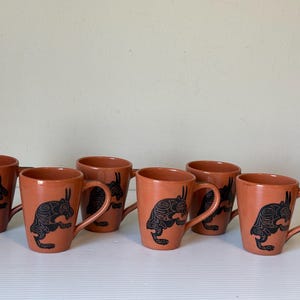Peut inclure: Ensemble de sept tasses en céramique couleur terre cuite. Chaque tasse présente un motif de lapin noir. Les tasses ont une forme cylindrique simple avec une anse incurvée. Les tasses sont présentées sur une surface blanche sur fond neutre.