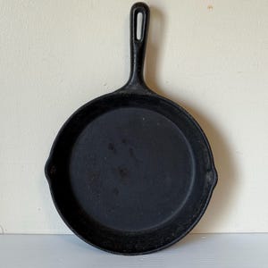 May include: A black cast iron skillet with a long handle and a round cooking surface. The skillet has a small pouring spout on either side of the rim. The handle has a hole for hanging. The skillet is designed for cooking.