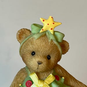 Puede incluir: Un oso de peluche marr&oacute;n vestido como un &aacute;rbol de Navidad. El oso lleva un vestido verde con una cinta amarilla y botones rojos. El oso tiene una estrella amarilla en la cabeza.