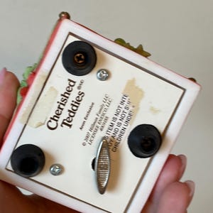 Puede incluir: Caja de m&uacute;sica blanca con un dise&ntilde;o floral rojo y verde. La caja tiene el logotipo de Cherished Teddies y una llave de cuerda en la parte inferior. El texto en la parte inferior dice "Este art&iacute;culo no est&aacute; destinado a ni&ntilde;os menores de 3 a&ntilde;os."