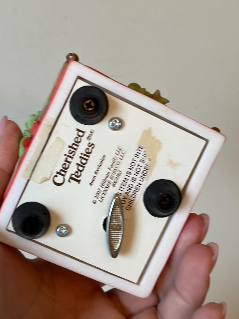 Puede incluir: Caja de m&uacute;sica blanca con un dise&ntilde;o floral rojo y verde. La caja tiene el logotipo de Cherished Teddies y una llave de cuerda en la parte inferior. El texto en la parte inferior dice "Este art&iacute;culo no est&aacute; destinado a ni&ntilde;os menores de 3 a&ntilde;os."