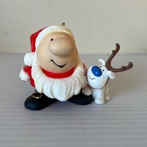 May include: A Christmas ornament featuring a white and red Santa Claus with a large nose and a white beard, holding a white reindeer with brown antlers.
