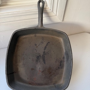 May include: A well-used, square cast iron skillet with a long handle and a hole for hanging. The pan is dark gray with signs of wear and use, indicating it's been seasoned. The handle is integrated into the pan's design.