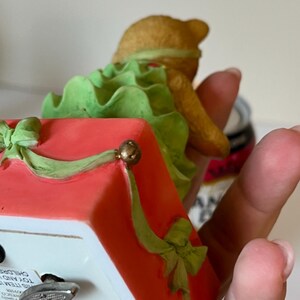 Puede incluir: Una figurita musical navide&ntilde;a de un oso de peluche con un vestido verde sentado en una caja roja con un lazo verde. La caja tiene una llave de cuerda y el texto "Cherished Teddies" en el lateral.