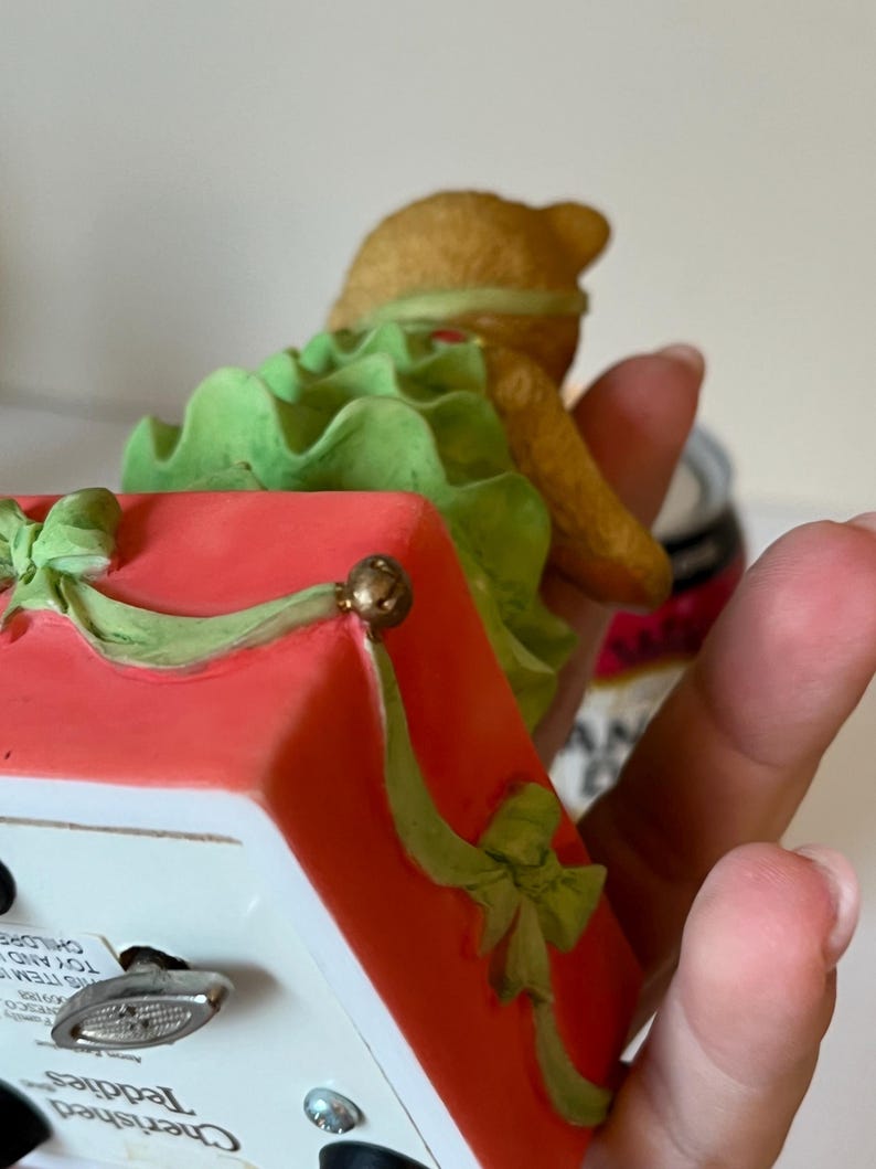 Puede incluir: Una figurita musical navide&ntilde;a de un oso de peluche con un vestido verde sentado en una caja roja con un lazo verde. La caja tiene una llave de cuerda y el texto "Cherished Teddies" en el lateral.