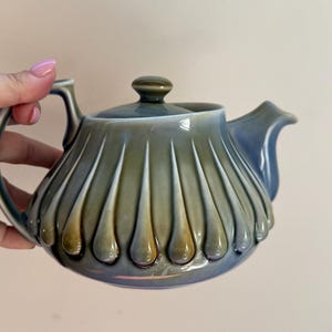 May include: A ceramic teapot featuring a rounded body with vertical fluted ridges and a glossy glaze that transitions from a muted olive green to a soft blue. The teapot includes a matching lid with a small knob and a curved handle and spout.
