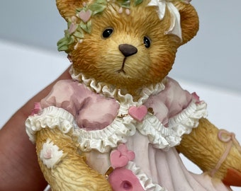 May include: A small, light brown teddy bear figurine wearing a pink dress with white lace trim and a floral crown. The bear is holding a pink heart-shaped object in its paw.