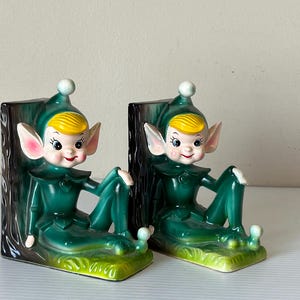 Puede incluir: Par de sujetalibros de cerámica vintage con forma de duende. Cada sujetalibros presenta un duende sentado con un atuendo verde, pelo amarillo y una cara sonriente. Los sujetalibros tienen un diseño de tronco de árbol y una base verde y amarilla.