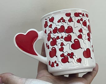 以下が含まれることがあります： 赤いハート型の取っ手が付いた白いセラミックマグ。マグカップは、白い背景に赤いハート型の風船で飾られています。バレンタインデーやあらゆる機会にぴったりの、魅力的でロマンチックなデザインです。