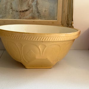May include: A yellow ceramic mixing bowl with a smooth white interior. The exterior features a decorative rope-like pattern along the rim and a series of embossed arch and diamond shapes around the body, resting on a square-shaped base.