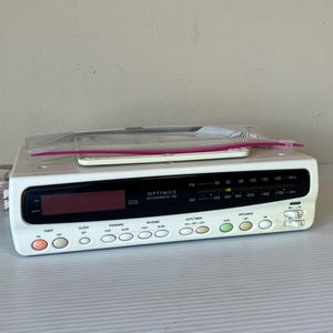 May include: White Optimus Kitchenmate 100 radio with a digital display and various function buttons. A clear plastic bag with a white remote is on top. The radio has a black panel with frequency indicators.