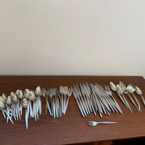 Puede incluir: Una gran colección de cubiertos de acero inoxidable en tono plateado dispuestos sobre una superficie de madera. El conjunto incluye numerosas cucharas, tenedores y cuchillos, junto con algunas piezas de servicio, con un diseño de mango texturizado.