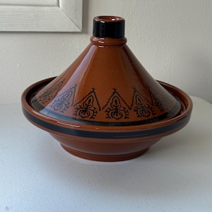 May include: A brown ceramic tagine pot with a conical lid and a wide base. The lid has black decorative patterns and a black handle. A black band encircles the base. This traditional cookware is designed for slow-cooked stews and tagines.