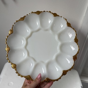 May include: A round white milk glass egg plate featuring twelve circular indentations arranged in a ring around a smooth center. The scalloped edge of the plate is finished with a metallic gold trim that highlights the curved rim design.