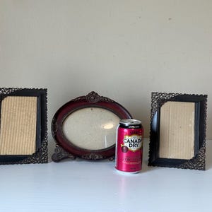 May include: Three antique picture frames are displayed. Two are rectangular with black frames and decorative corner patterns. The third is oval with a burgundy frame. A can of Canada Dry is in the center.
