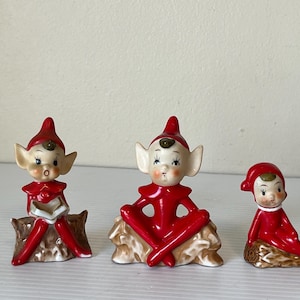 Puede incluir: Tres figuras de elfos de cerámica con trajes y sombreros rojos. Un elfo lee un libro, otro está sentado con las piernas cruzadas y el tercero está sentado con las piernas extendidas. Las figuras están sobre bases marrones.