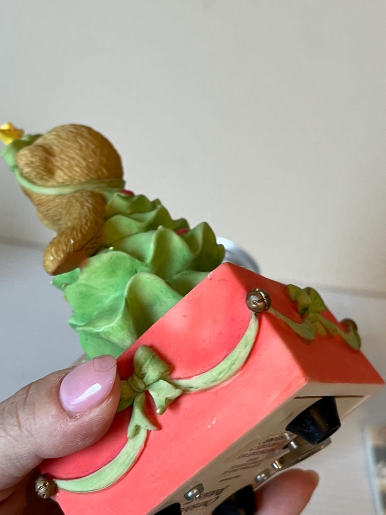 Puede incluir: Una figurita musical de &aacute;rbol de Navidad con un oso marr&oacute;n vestido como &aacute;rbol de Navidad. La figurita est&aacute; en una caja roja con una cinta verde y una campana dorada.