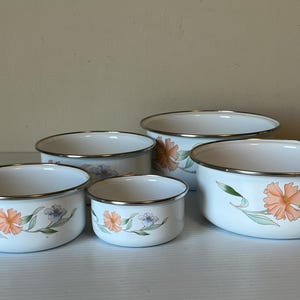 May include: A set of five white enamel bowls with a floral design. Each bowl features a silver rim and is decorated with orange and purple flowers and green leaves. The bowls vary in size.