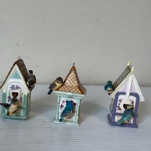 Könnte beinhalten: Drei dekorative Vogelhäuser in Pastellfarben, jedes mit einem anderen Vogel verziert. Die Häuser weisen aufwendige architektonische Details und florale Muster auf und erzeugen ein charmantes und skurriles Aussehen. Dekorative Objekte.
