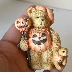 Puede incluir: Un par de osos de peluche vestidos con disfraces de calabaza para Halloween. El oso más grande sostiene una calabaza tallada en un palo.