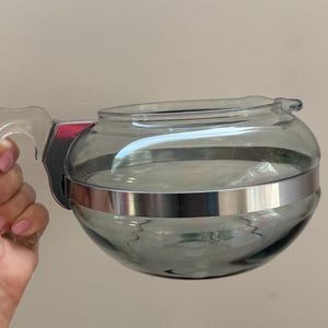 May include: A round, smoke-grey glass coffee carafe featuring a wide, polished silver-tone metal band encircling the centre. The vessel includes a clear, curved glass handle attached to the body with a silver-tone metal bracket and a small spout.