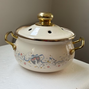 Op de afbeelding: Een crèmekleurige emaille pan met een goudkleurige dekselknop en twee handgrepen aan de zijkant. De pan is voorzien van een decoratief patroon van witte ganzen, blauwe linten en kleine rode appels. Het deksel heeft diverse kleine ventilatiegaten.
