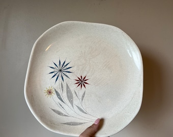 May include: A white ceramic dinner plate with a floral design. The design features blue, red, and yellow flowers with gray stems and leaves.