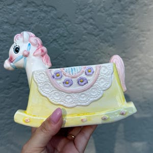 May include: A ceramic rocking horse figurine featuring a white horse head with pink hair and a blue bridle. The body is a yellow rocker base decorated with a white lace-like saddle pattern, accented by small purple flowers and pink trim details.