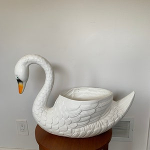 May include: A large, white swan planter with a detailed feather design. The swan has a long, curved neck and an open basin for plants. The beak is orange and the eyes are green. The planter is sitting on a small, round, wooden table.