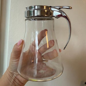 May include: A clear glass syrup dispenser featuring a polished chrome metal lid and a matching curved handle. The top mechanism includes a thumb-operated lever designed for controlled pouring, with a wide base and a tapered neck structure.