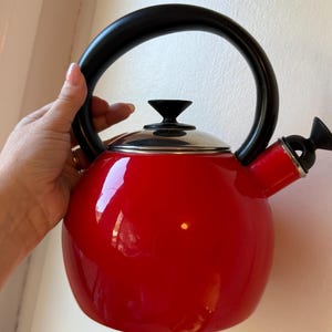 May include: A bright red tea kettle with a glossy finish, featuring a rounded body, a black arched handle, a black lid knob, and a black spout cap. The kettle has a metallic silver lid base and a matching silver ring around the spout opening.