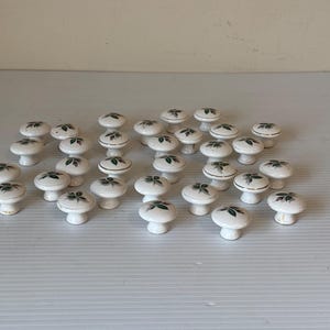 Peut inclure: Ensemble de boutons de meubles en céramique blanche avec un motif floral. Chaque bouton a une base blanche, un dessus arrondi avec un motif de feuilles vertes et une bordure dorée. Les boutons sont disposés sur une surface blanche.