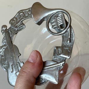 Puede incluir: Plato de metal plateado con un diseño de instrumentos musicales, incluyendo un violín, una trompeta y notas musicales. El plato es de vidrio transparente y se encuentra sobre una superficie blanca.