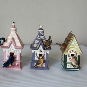 Könnte beinhalten: Drei dekorative Vogelhäuser in Pastellfarben. Die Vogelhäuser sind rosa, lila und grün, jedes mit einem anderen Design und kleinen Vogelfiguren, die darauf sitzen. Die Vogelhäuser haben herzförmige Öffnungen.