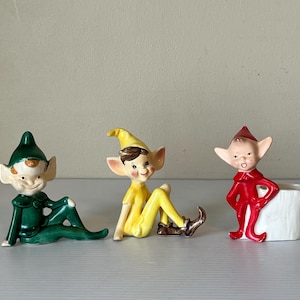 May include: Three ceramic elf figurines in green, yellow, and red. The green elf is reclining, the yellow elf is seated, and the red elf stands next to a white planter. Each elf has a pointed hat and large ears.