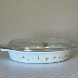 Puede incluir: Una fuente para horno Pyrex blanca vintage dividida con tapa de cristal transparente. El plato presenta un patrón de estrellas doradas. El plato ovalado tiene asas, ideal para servir. Mide aproximadamente 25 cm de largo.