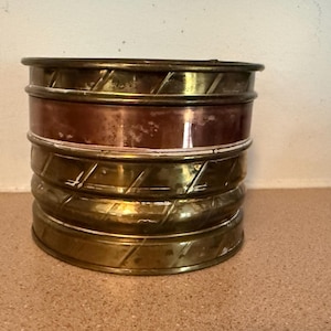May include: A stack of four cylindrical metal containers. The top, third, and bottom containers are made of brass with a diagonal ribbed pattern. The second container from the top is made of smooth, polished copper with visible surface oxidation.