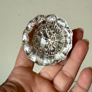 Puede incluir: Un pomo de puerta de cristal transparente y facetado con un diseño circular. El pomo tiene una superficie detallada y texturizada y un patrón central en forma de estrella. El cristal refleja la luz, creando un efecto brillante.