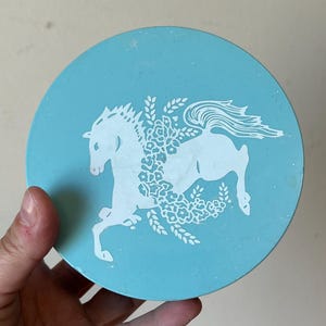 May include: A light blue round tin with a white horse design. The horse is surrounded by white flowers and leaves.