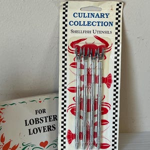Op de afbeelding: Een verpakking van Culinary Collection schaaldierbestek met vier zilveren vorken. De verpakking heeft een zwart-wit geblokte rand en rode krabillustraties. Tekst op de verpakking luidt "Shellfish Utensils" en "How to Eat and Prepare Seafood".
