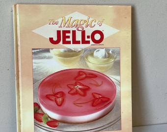 Op de afbeelding: Een vintage kookboek getiteld "The Magic of Jell-O" met een dessertafbeelding op de omslag. De omslag toont een rood en wit gelaagd dessert met aardbeienschijfjes. Het boek viert 100 jaar Jell-O-recepten.