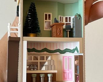 May include: A miniature replica of a vintage soda fountain with a pink door, a green awning, and a sign that reads "YUMMY SODA FOUNTAIN". The interior features a counter with stools, a small Christmas tree, and a desk with a chair.