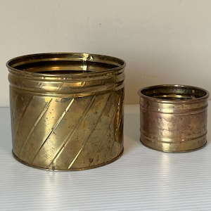Peut inclure: Deux récipients en laiton antiques. Le plus grand présente un motif nervuré en diagonale, tandis que le plus petit a une texture martelée. Les deux sont cylindriques et ont une teinte dorée chaude, suggérant l'âge et l'utilisation.