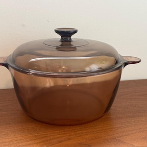 Op de afbeelding: Een ronde, doorschijnende amberkleurige glazen ovenschaal met twee zijhandgrepen en een bijpassend glazen deksel met een centrale knop. Het kookgerei heeft een gladde, gepolijste afwerking en een brede, diepe komvorm voor bereiding of opdiening.
