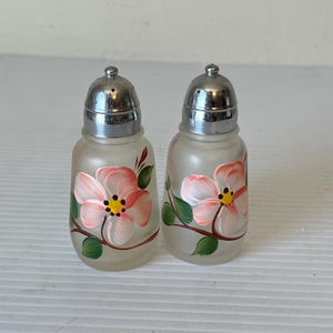 May include: A pair of frosted glass salt and pepper shakers with silver metal lids. Each shaker features a hand-painted pink flower with yellow centres and green leaves. The shakers are a vintage design.