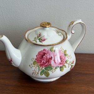 Puede incluir: Una tetera de porcelana blanca con un estampado floral de rosas rosas y hojas verdes. La tetera tiene una tapa festoneada con un pomo dorado, un asa curva con detalles en oro y un borde dorado alrededor de la tapa y la abertura del pico.