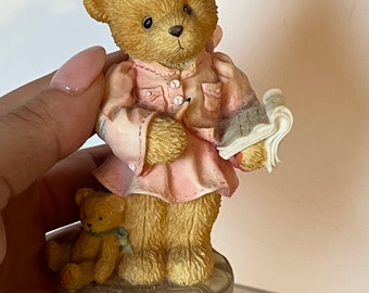 May include: A figurine of a brown teddy bear wearing a pink dress and holding a book and pencil. The bear is standing on a round base with a smaller brown teddy bear sitting beside it.
