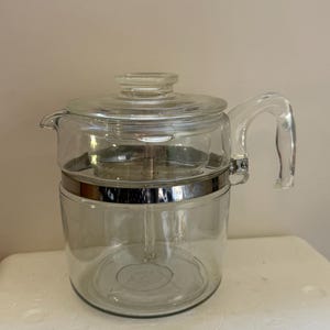 Puede incluir: Una cafetera percoladora de vidrio transparente con tapa, un asa de vidrio resistente y una banda de metal plateado que rodea el centro. El diseño incluye un tubo central de vidrio y una cesta de vidrio para la preparación del café.