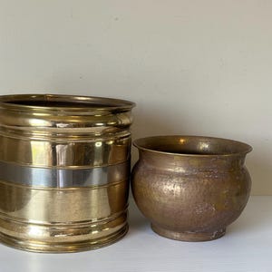 May include: Two brass planters. One is a cylindrical shape with a silver band around the middle. The other is a smaller, hammered-texture bowl shape. Both are on a white surface.