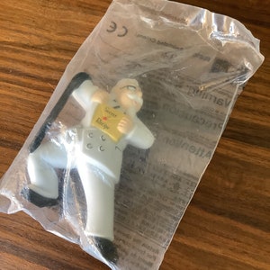 Puede incluir: Una figurita de plástico blanco de un chef que sostiene un sobre amarillo con las palabras "Secret Recipe" en él. El chef lleva un gorro de cocinero blanco y una chaqueta blanca con botones negros. La figurita está empaquetada en una bolsa de plástico transparente.